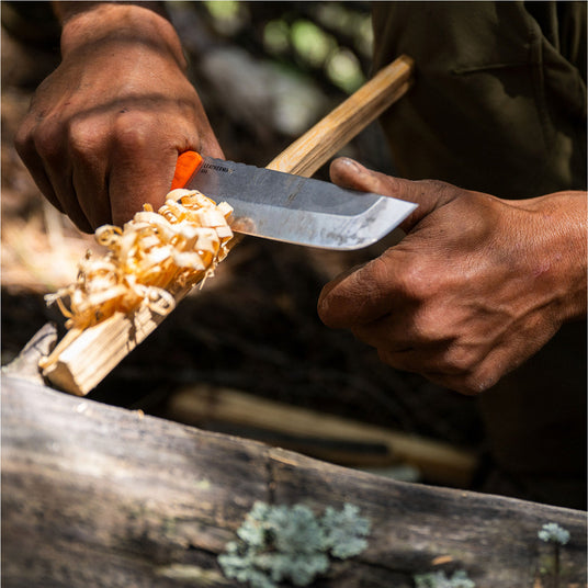 Translation missing: fr.Person using a Leatherman Pioneer knife to carve feather sticks from a wooden branch in an outdoor setting, shown in a close-up highlighting controlled bushcraft technique