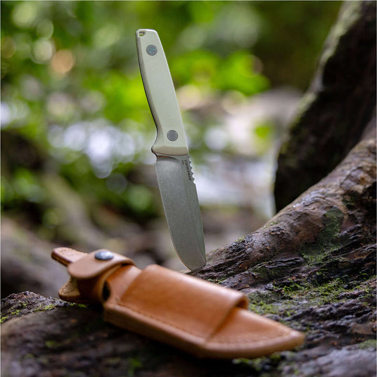 Translation missing: fr.A Leatherman Trac fixed-blade knife is vertically embedded in a mossy tree trunk in a lush forest setting. The knife features a light-colored handle with visible fasteners, and a leather sheath lies in the foreground on the log