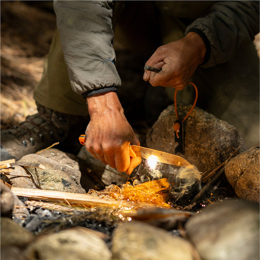 Translation missing: fr.Person using a Leatherman Pioneer knife and fire starter to ignite feather sticks in a campfire setup, shown in an outdoor setting with sparks and smoke rising from the tinder