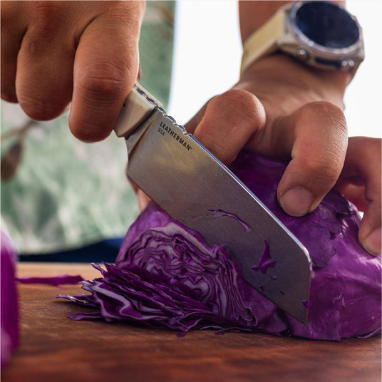 Translation missing: en.Person using a Leatherman Rustle knife to slice a head of purple cabbage on a wooden cutting board, shown in a close-up with focus on the blade and hands in action