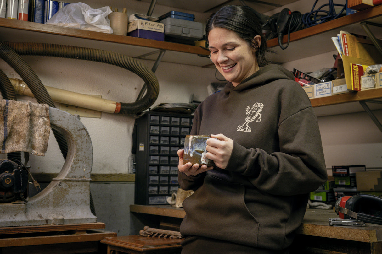 Translation missing: fr.A person wearing a brown Leatherman Mr. Wave hoodie standing in a workshop holding a ceramic mug, smiling while surrounded by tools and equipment