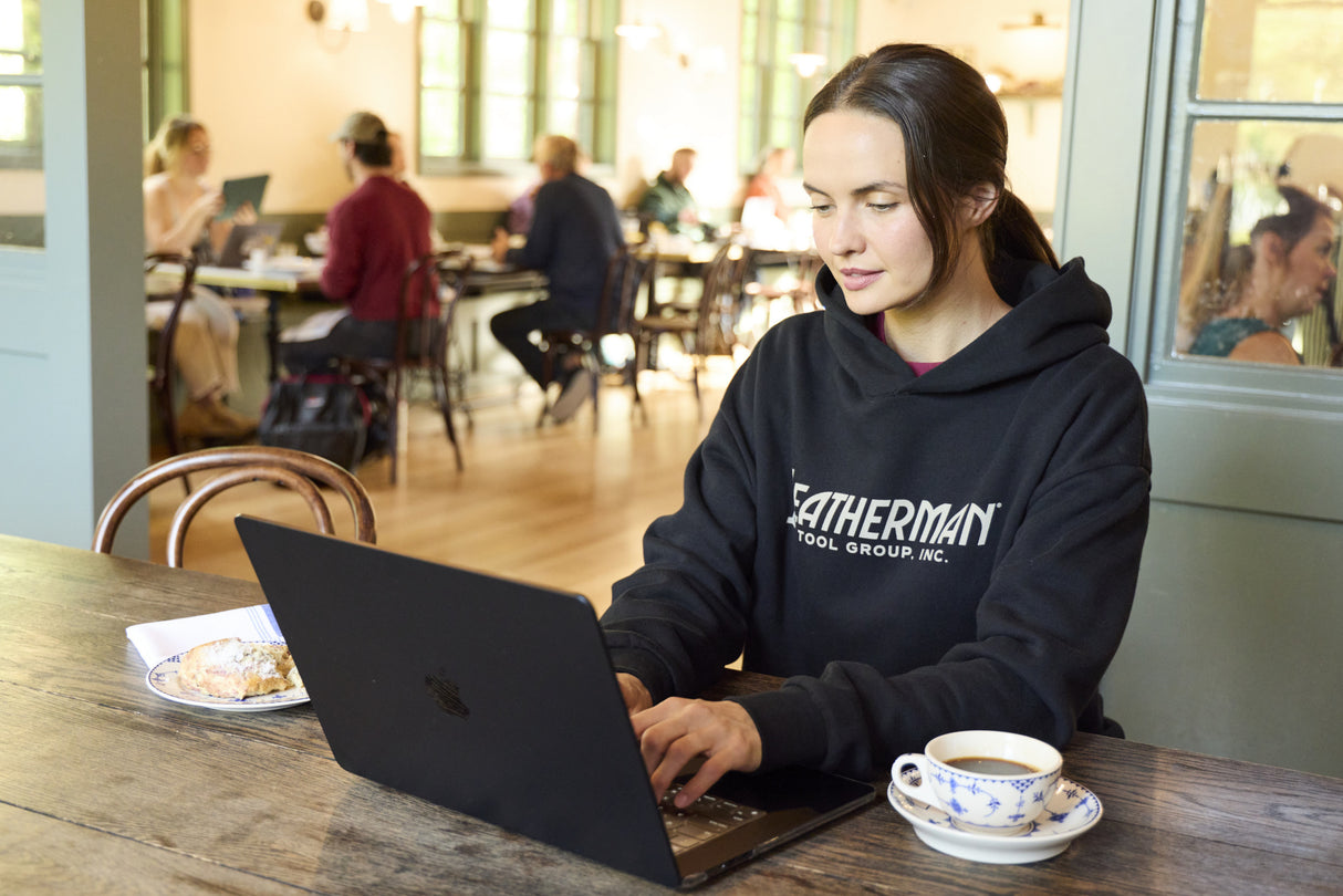 Translation missing: fr.A woman sitting indoors working on a computer while wearing a black Leatherman Blade & Grain hoodie with a pastry and coffee on either side of the laptop