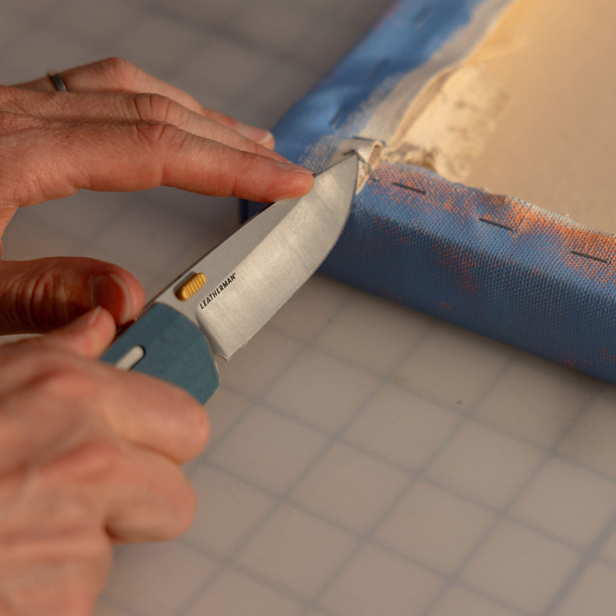 Translation missing: en.Person using a Leatherman Blazer Denim folding knife to cut canvas material from a wooden frame, shown in a close-up on a gridded surface