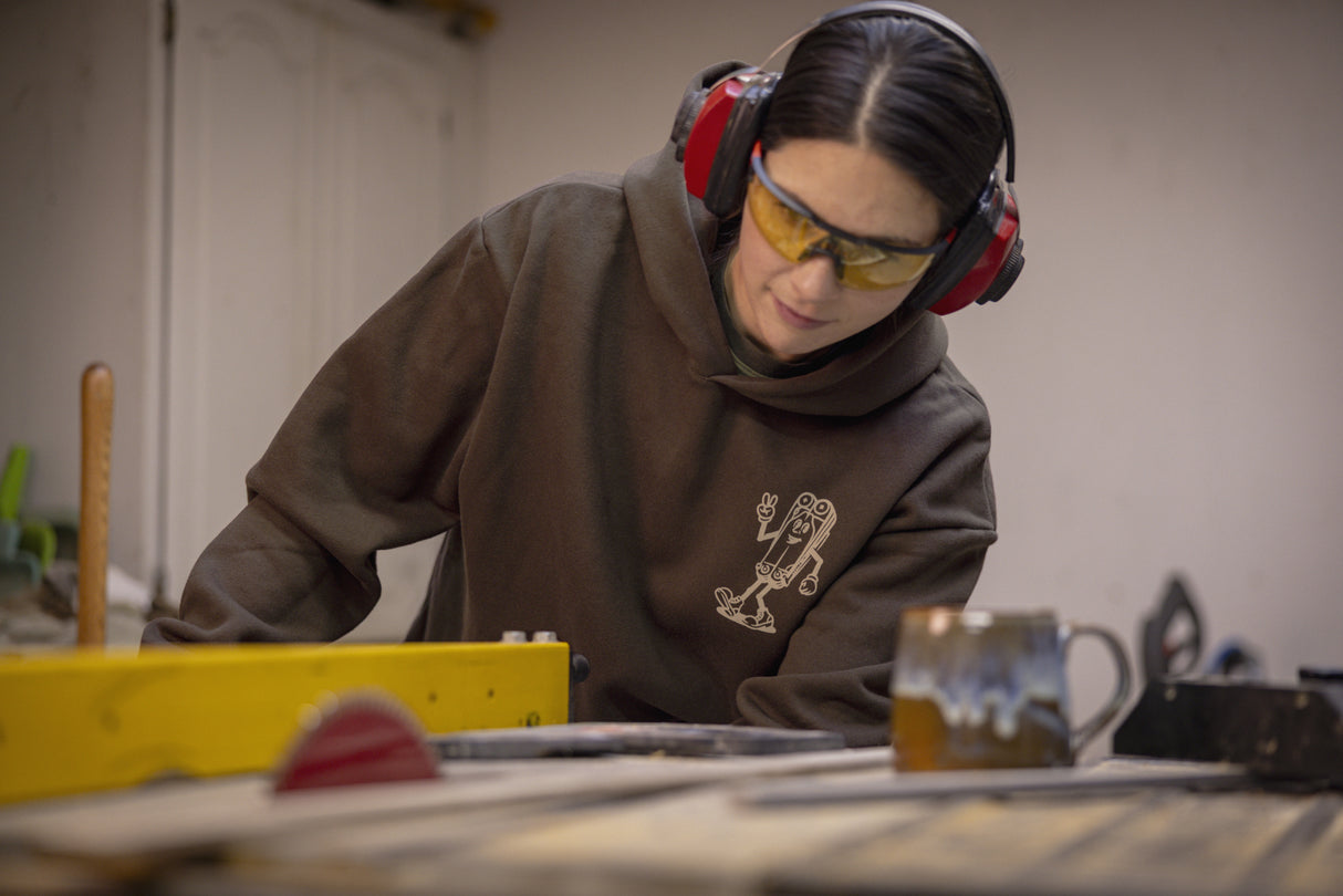 Translation missing: fr.A person wearing a brown Leatherman Mr. Wave hoodie operating a table saw in a workshop, wearing safety glasses and earmuffs for protection