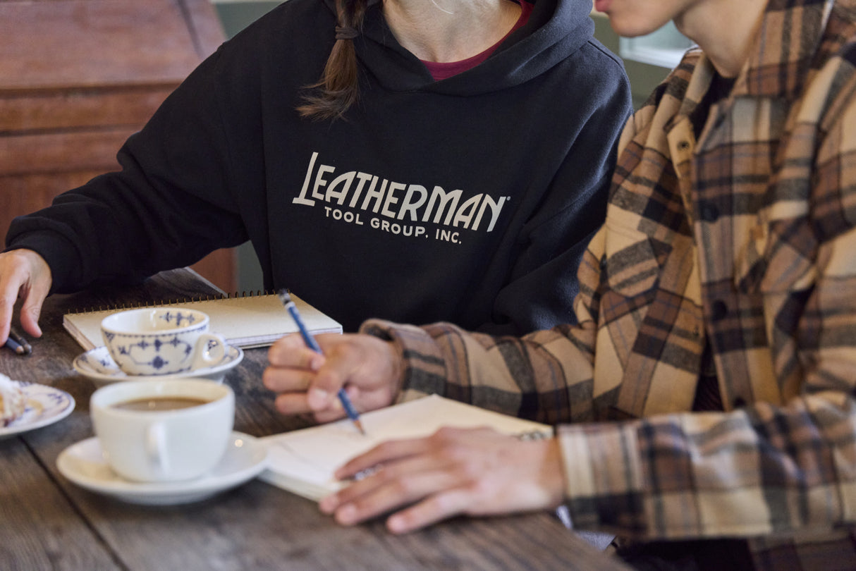 Translation missing: fr.Two people sitting at a table with coffee, pastries and notebooks. One is wearing a black Leatherman Blade & Grain hoodie with 'Leatherman Tool Group Inc.' on the front