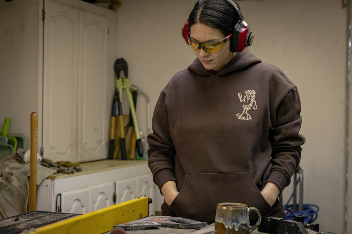 Translation missing: fr.A person wearing a brown Leatherman Mr. Wave hoodie standing in a workshop with safety glasses and ear protection, looking down at a workbench with tools and a ceramic mug