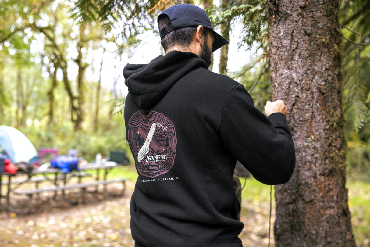 Translation missing: fr.A person wearing a black Leatherman Blade & Grain hoodie standing next to a tree in a forest campsite. The hoodie’s back graphic shows a pocketknife illustration with “Leatherman Tool Group” text