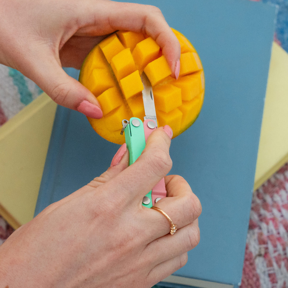 Translation missing: fr.Hands using a Leatherman Micra® in Bayside color to cut a mango on top of a stack of books