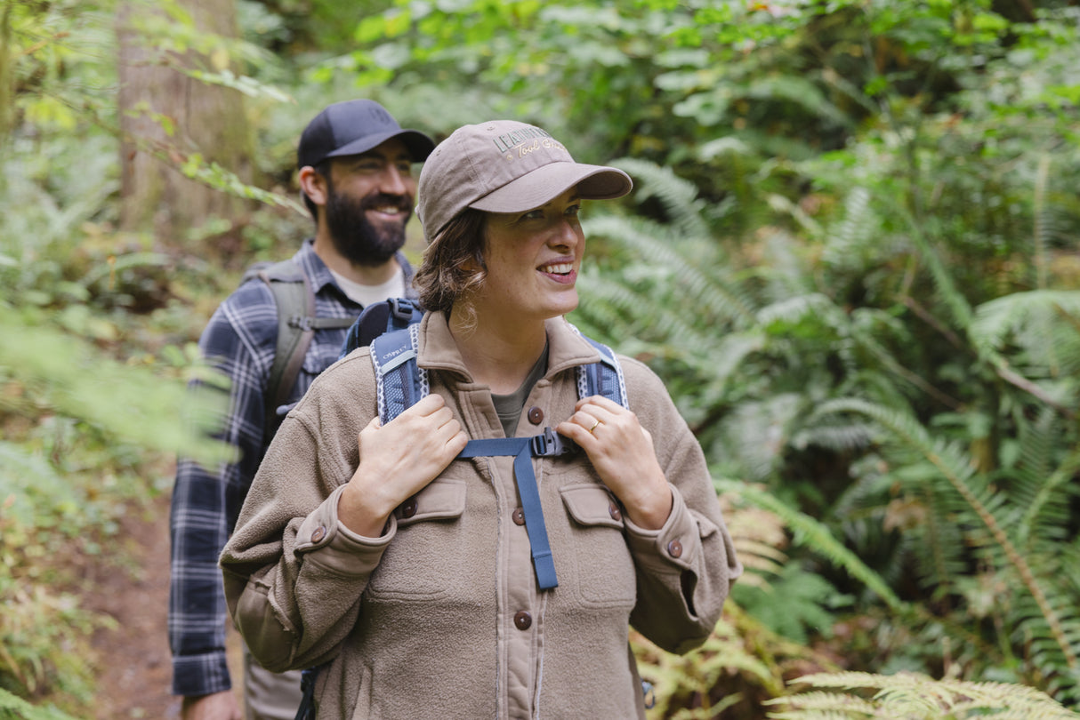Translation missing: fr.A woman wearing a Leatherman Dad Hat in Driftwood color hiking through a forest with a backpack smiling as another hiker follows behind