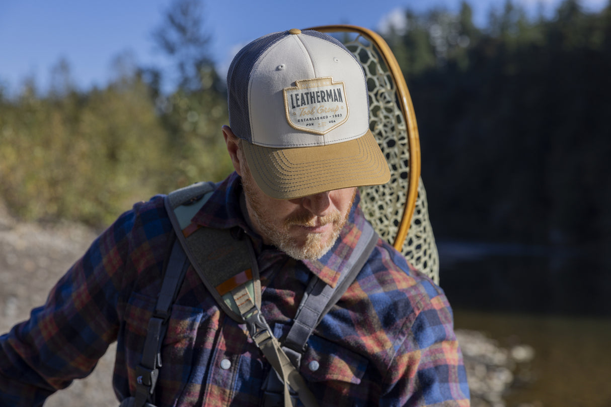 Translation missing: fr.A person wearing a tan and brown Leatherman PNW Trucker Hat standing near a river with a backpack and fishing net