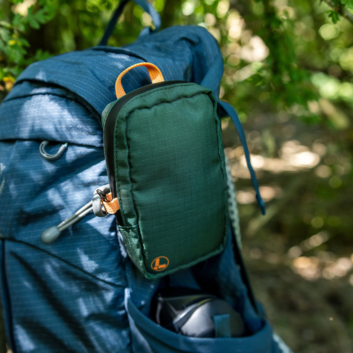 Translation missing: fr.A Leatherman Tool Pouch in Hunter color is clipped to the shoulder strap of a blue hiking backpack, resting in an outdoor forest setting with sunlight filtering through the trees