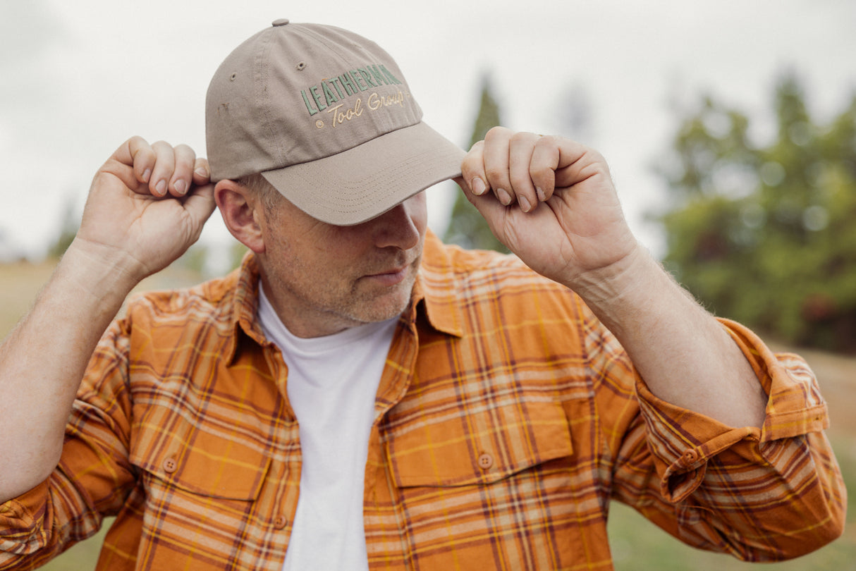 Translation missing: fr.A person wearing a Leatherman Dad Hat in Driftwood color adjusting the brim while outdoors, dressed in an orange plaid shirt