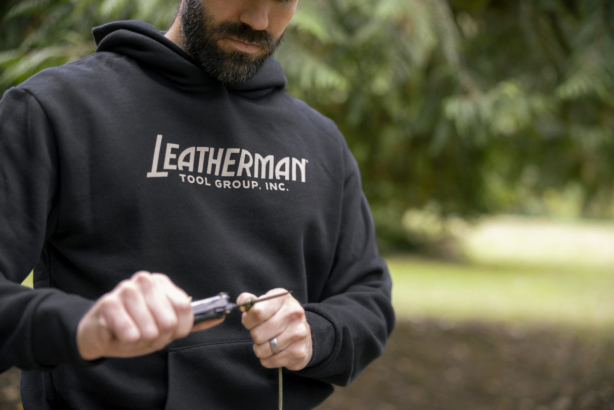 Translation missing: fr.A person wearing a black Leatherman Blade & Grain hoodie using a Leatherman multitool to cut a piece of rope outdoors, showing the front logo of the hoodie that reads “Leatherman Tool Group, Inc.”