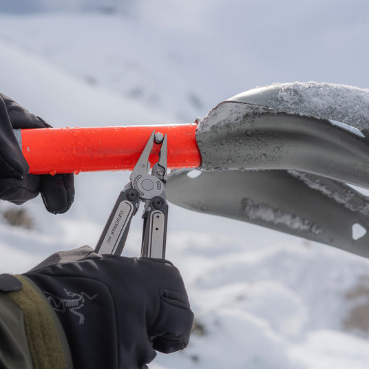 Translation missing: fr.Person wearing black gloves using a Leatherman Arc multi-tool to tighten a bolt on an orange-handled snow shovel in a snowy mountain environment