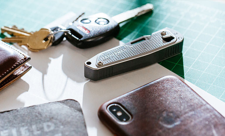 Translation missing: fr.Brown Leatherman Wallet, car keys, Leatherman T2, gray note pad, and Iphone on top of white piece of paper placed on green grid table