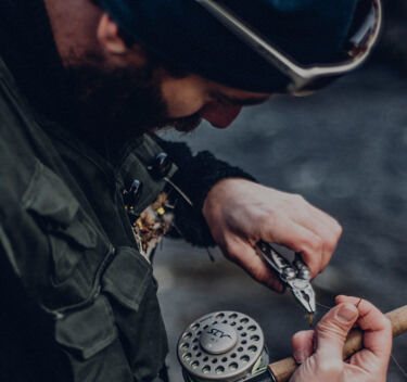 Translation missing: fr.A man using Leatherman Skeletool to tie a fly to his fishing rod, demonstrating his preparation for fly fishing