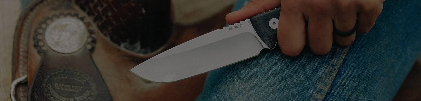Translation missing: en.Close-up of a person holding a Leatherman fixed-blade knife with "MAGNACUT" engraved on the blade, positioned over their denim-clad leg next to a leather saddle