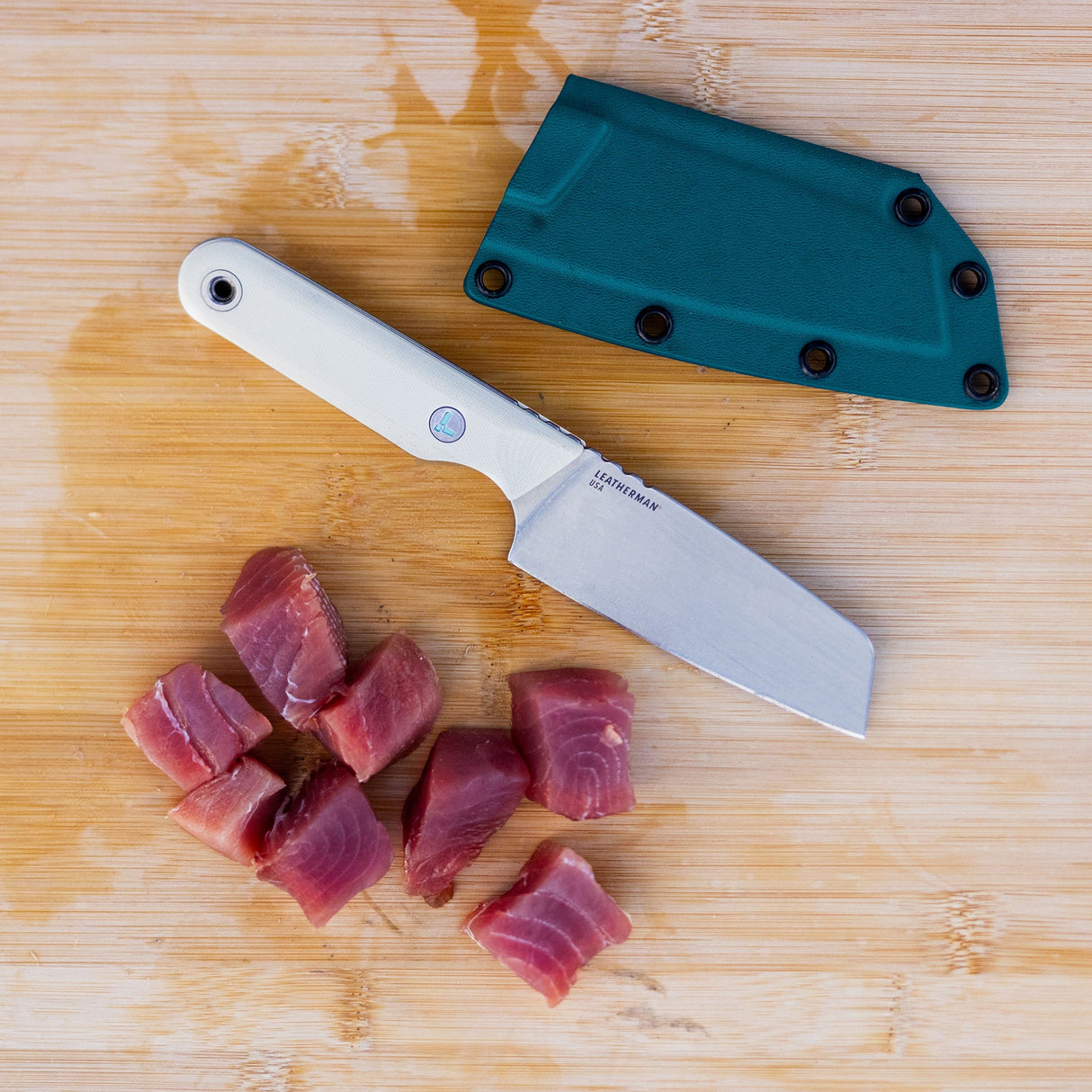 Translation missing: en.A Leatherman Rustle Alpine knife on a wooden cutting board next to a teal sheath and freshly cut chunks of raw fish