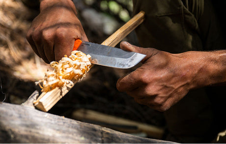 Translation missing: fr.Close-up of a person using a Leatherman knife with an orange handle to carve wood shavings from a stick, surrounded by natural outdoor elements, suggesting a bushcraft or survival setting