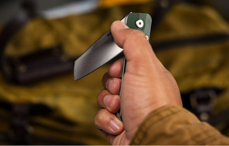 Translation missing: fr.Close-up of a person holding a partially open Leatherman folding knife with a green handle, positioned in front of a yellow canvas bag with leather straps