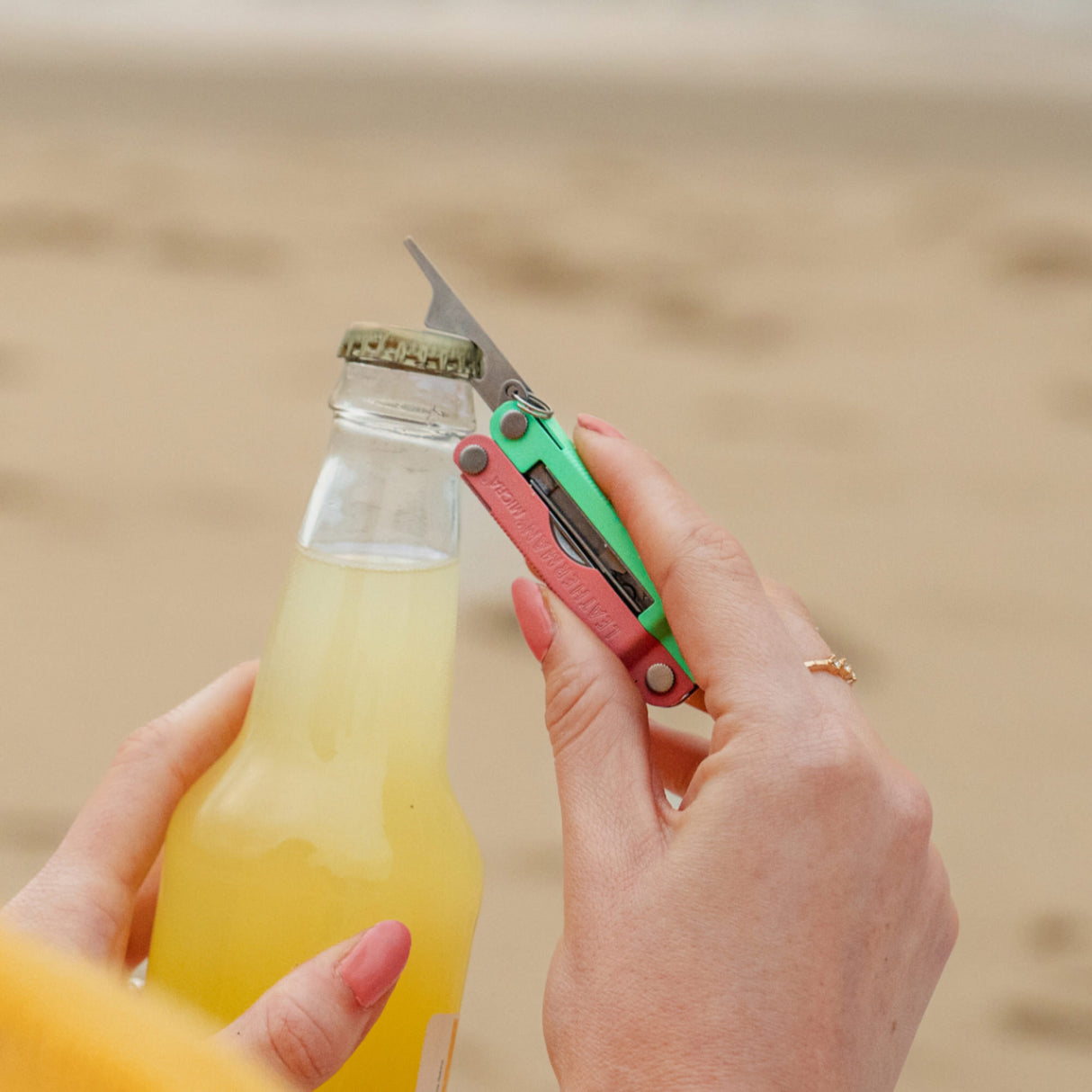 Translation missing: fr.Person using the bottle opener on a Leatherman Micra® in Bayside color to open a bottle on the beach