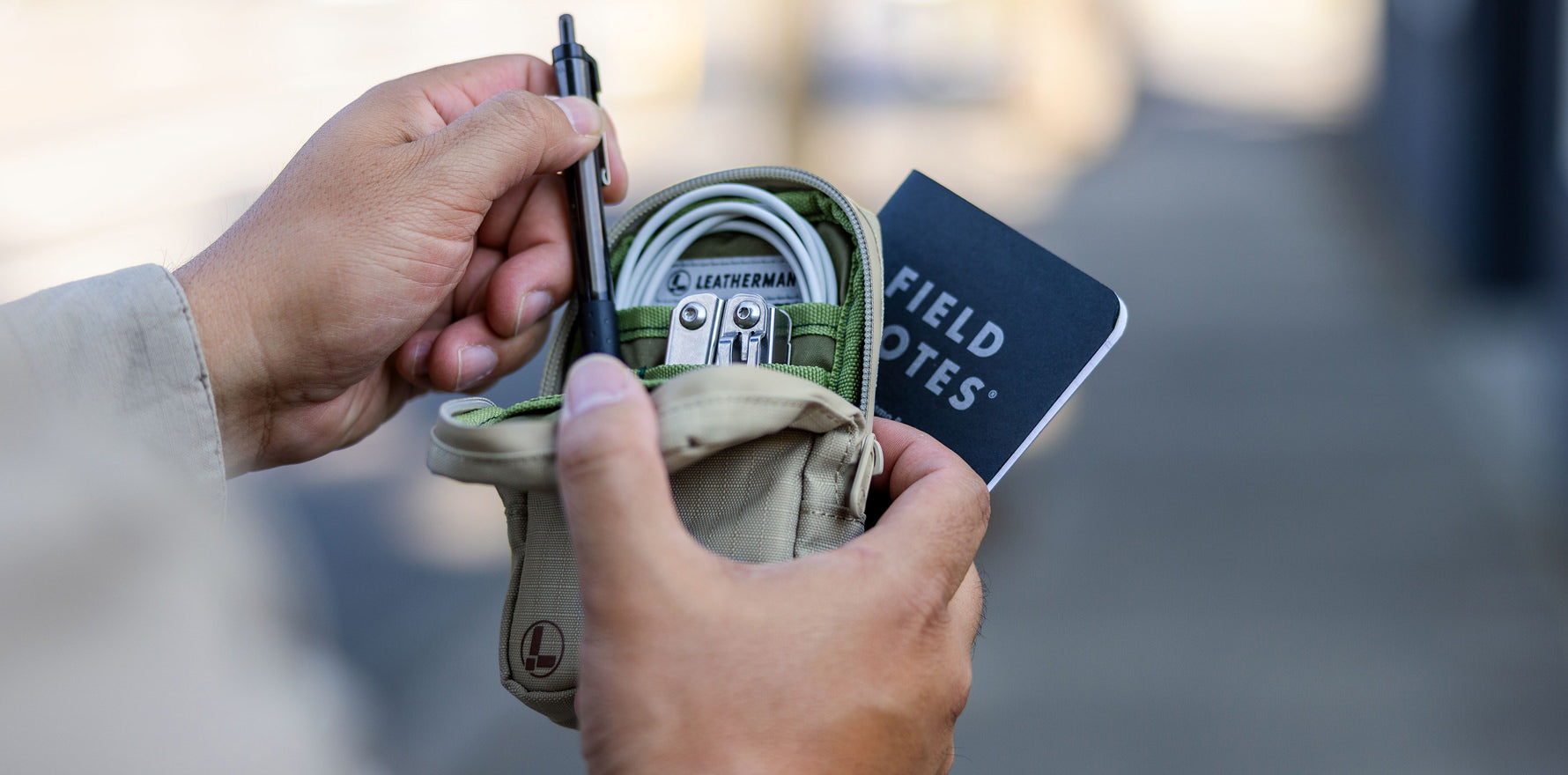 A person holds open a Leatherman Tool Pouch in Mossy Fog color, revealing neatly organized items including a Leatherman multi-tool, charging cable, pen, and Field Notes notebook—illustrating the pouch's utility for everyday carry and organization.