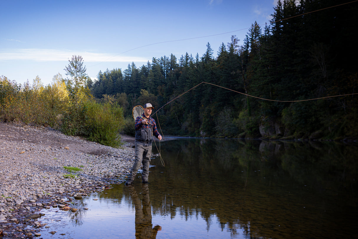 Translation missing: en.A person wearing a beige Leatherman PNW Trucker hat fly-fishing in a river surrounded by trees