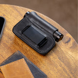 Translation missing: en.Black leather sheath with belt clip holding a small cylindrical flashlight on a wooden surface