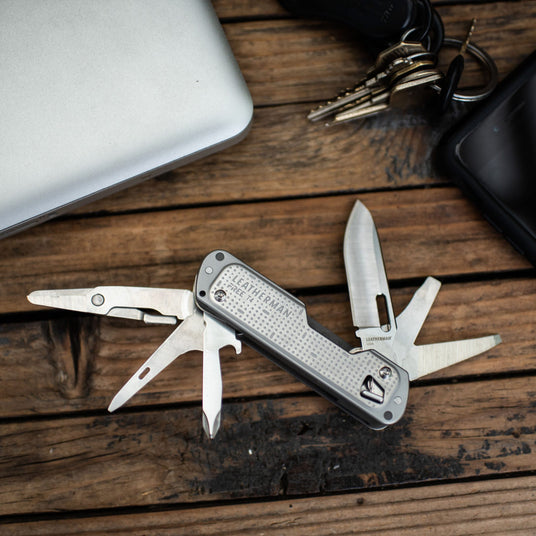 FREE T4 with knife, and other tools extended, lying next to a laptop, keys, and smartphone on a wooden surface.