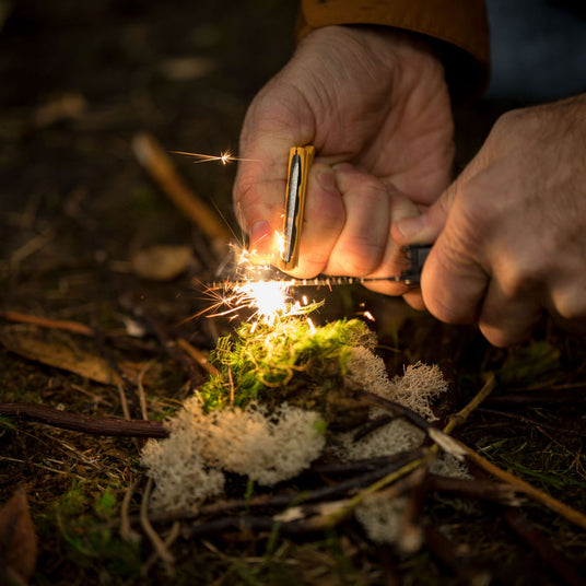 Close-up hands using a Leatherman Signal to start a fire in the wilderness with a fire-starting ferro rod.