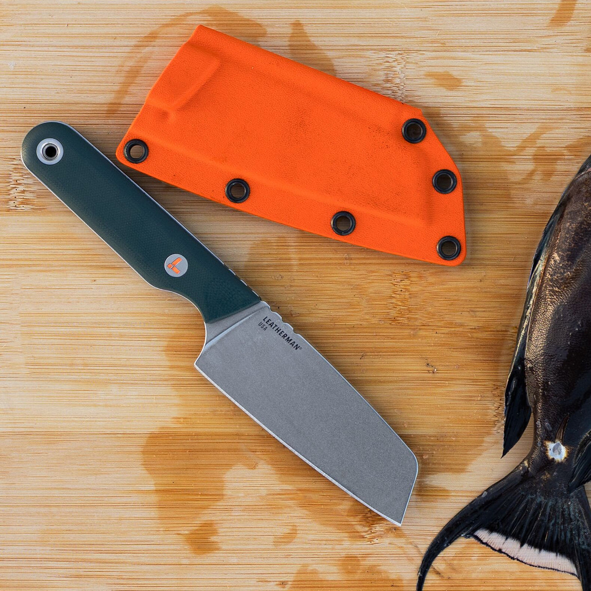 Translation missing: en.Leatherman Rustle Evergreen knife with a green handle and stainless steel blade resting on a wooden cutting board next to its bright orange sheath and a partially cleaned fish