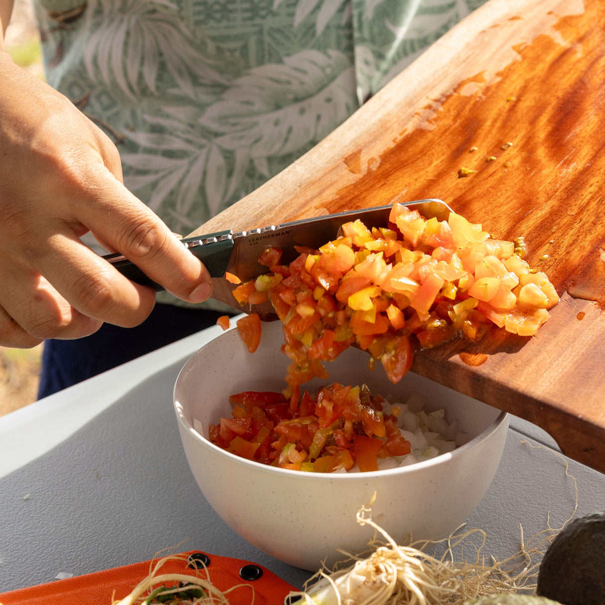 Translation missing: en.Person using a Leatherman Rustle Evergreen knife with a green handle to transfer freshly chopped tomatoes and vegetables from a wooden cutting board into a white bowl, outdoors on a sunny day
