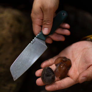 Translation missing: en.Person holding a Leatherman Rustle Evergreen knife with a green handle in one hand and dried dates in the other, suggesting outdoor food preparation or foraging