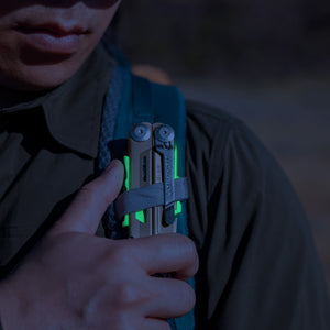 Translation missing: en.Person securing a Sandstorm multitool with neon accents to a backpack strap outdoors
