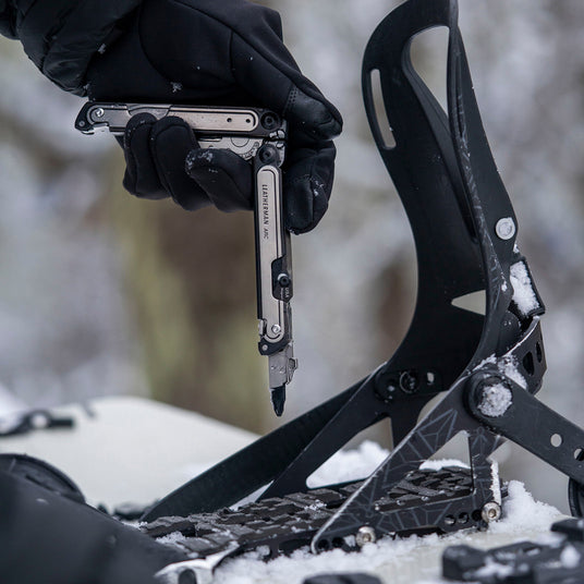 Translation missing: en.Person using Black DLC &amp; Stainless Steel Leatherman Arc® multitool while adjusting snowboard bindings in the snow